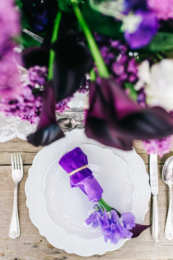 Close-up detail shot of a wedding reception place setting on a rustic wooden table. A white scallop-edged charger plate holds a deep violet linen napkin rolled and tied with twine, with a sprig of lavender sweet peas tucked into the knot. Silver vintage-style cutlery — a fork to the left and a knife and spoon to the right — flank the plate. The background is softly blurred but shows a lush centerpiece arrangement of deep burgundy-black calla lilies, purple hydrangeas, lilac blooms, and white flowers with green foliage, alongside a piece of white lace fabric. The overall decor palette is deep purple, violet, and white with a rustic-romantic styling theme.