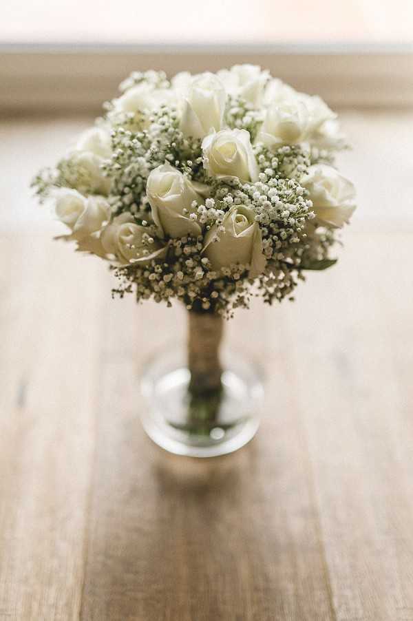 Close-up detail shot of a bridal bouquet resting in a small clear glass vase on a light wood surface. The compact, rounded bouquet is composed of white roses and abundant baby's breath, with the stems wrapped together. Natural window light illuminates the arrangement from behind, creating a soft, bright background. The styling is classic and minimal, with an all-white floral palette.