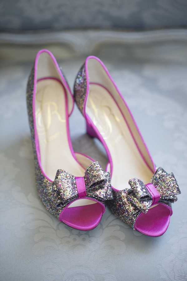 Close-up detail shot of a pair of bridal shoes placed on a gray damask-patterned surface. The heels are peep-toe pumps covered in multicolored chunky glitter with magenta pink satin trim, peep-toe binding, and a glitter bow detail at the toe. The insoles are cream-colored with a designer logo embossed in gold. The bold magenta and glitter combination reflects a playful, fashion-forward bridal styling choice.