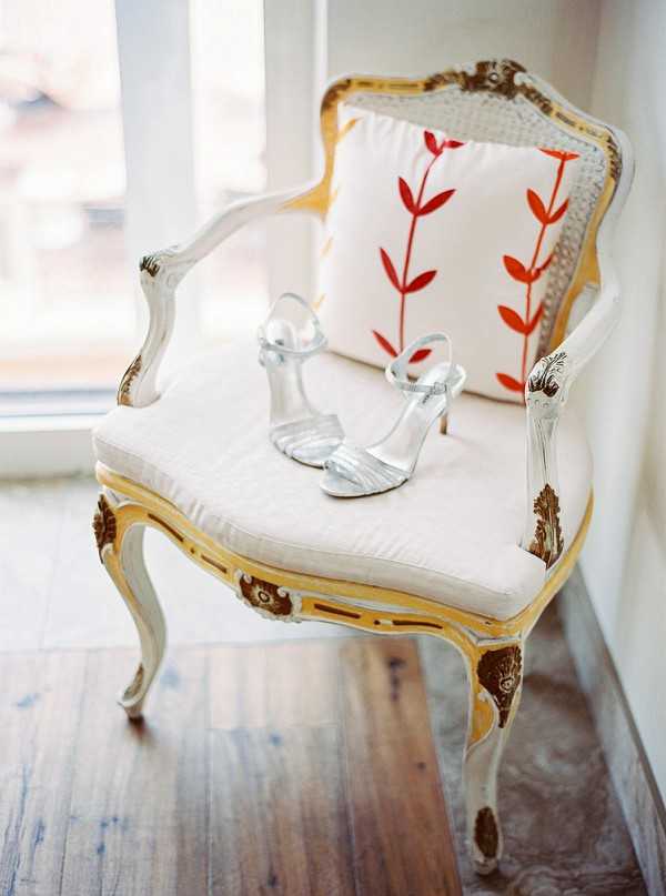 A detail close-up shot of a pair of silver strappy heeled sandals placed on the seat of an ornate French Louis XV-style armchair with a distressed white and gold painted frame, carved wooden details, and a cream upholstered cushion. A decorative throw pillow with a red botanical leaf print leans against the chair's back. The chair sits on a worn hardwood floor near a bright window, lending soft natural light to the scene. The styling reflects a classic French vintage aesthetic.
