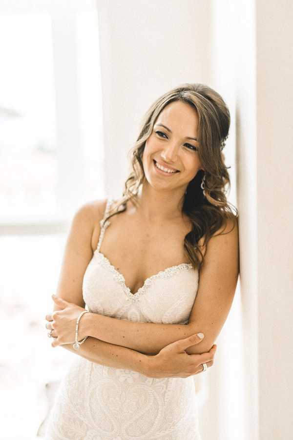 A bridal portrait of a smiling bride leaning against a white wall or door frame in a bright, naturally lit indoor setting, likely during a getting-ready moment. She wears a fitted ivory lace gown with a sweetheart neckline and thin beaded straps, paired with a delicate diamond tennis bracelet, a ring, and drop earrings. Her dark brunette hair is styled in loose waves swept to one side, and she has her arms crossed lightly in front of her. The composition is a close-up portrait shot with soft, bright backlighting from a nearby window creating a light, airy feel.