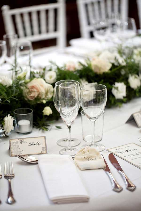 Close-up detail shot of a wedding reception table setting with a white linen tablecloth and white chiavari chairs visible in the background. The place setting includes two empty wine glasses, silver flatware, a folded white napkin with a calligraphed 'Merci' tag, a handwritten place card reading 'Kathleen' with a decorative botanical border, and a printed menu card. A small silver votive candle holder sits nearby. The centerpiece running along the table features a lush garland of green foliage interspersed with blush garden roses and ivory white ranunculus or lisianthus blooms, creating a classic, refined table design in a white and blush palette.