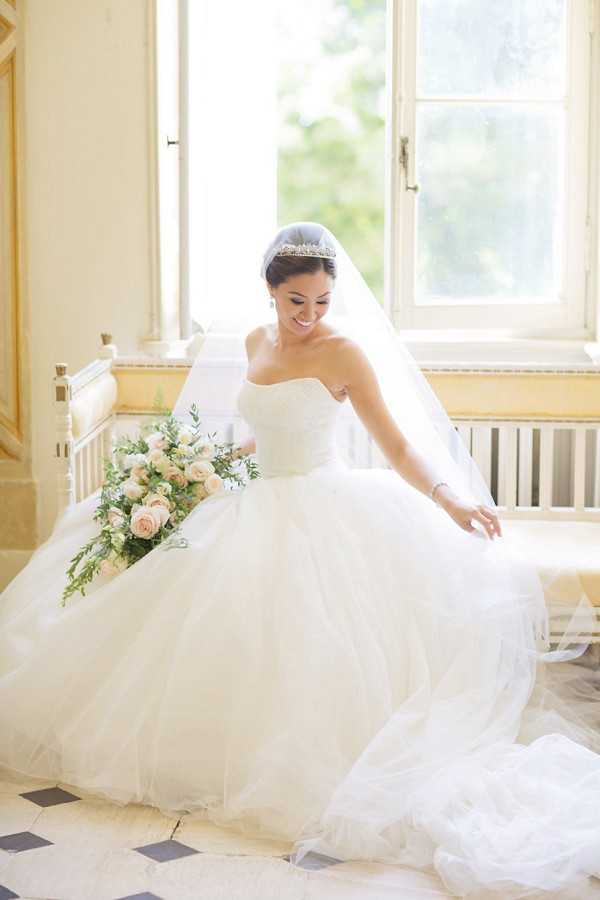 A bridal portrait of a bride seated indoors in what appears to be a chateau or historic French venue, shot in a medium portrait composition with soft natural window light. The bride wears a strapless white ball gown with a voluminous layered tulle skirt, a crystal tiara, and a cathedral-length veil, and she is looking down with a smile while arranging her skirt. She holds a loosely arranged bouquet of blush roses and green foliage. The interior setting features warm yellow-toned walls, a black-and-white tiled floor, and tall French windows, consistent with a classic chateau aesthetic.