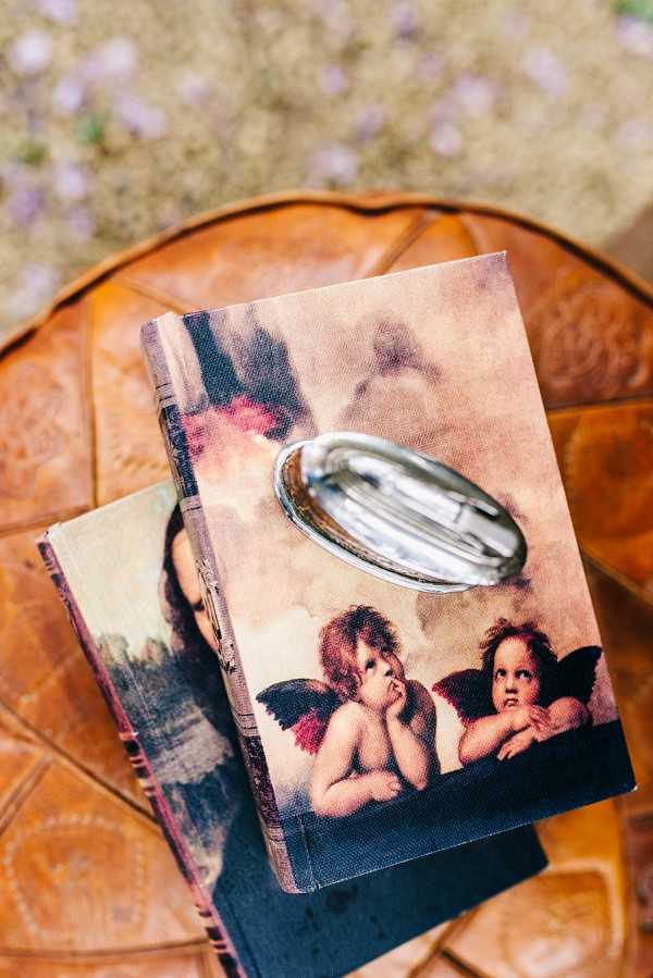 A close-up detail shot of two stacked decorative books featuring Raphael's cherub print on their covers, placed on a warm brown leather surface. A silver oval ring dish or cufflink holder sits on top of the upper book, holding what appear to be wedding rings or small metal accessories. The books have a vintage, art-inspired aesthetic with the classic Renaissance cherub imagery in muted rose and warm beige tones. Small blurred pale purple florals are visible in the soft-focus background.