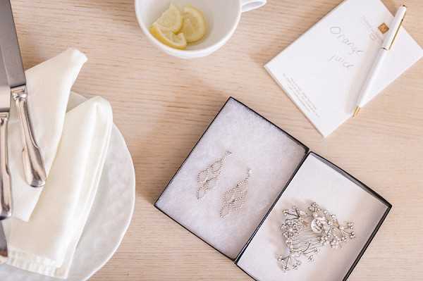 A flat lay detail shot taken from above on a light wood surface, showing bridal jewelry laid out in an open black gift box with white fabric lining. The box contains a pair of silver crystal drop earrings with a feather-like design and a matching silver crystal brooch or hair accessory. Also visible are a white folded napkin on a plate with silverware, a white cup containing lemon slices, and a small white stationery card with gold script lettering reading 'Orange Juice' alongside a gold and white pen. The overall styling palette is white and silver with clean, minimal presentation.