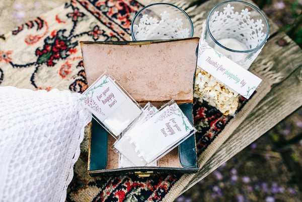 A close-up flat lay detail shot of wedding favors and ceremony accessories arranged on a patterned vintage rug draped over a wooden surface. An open vintage teal jewelry or keepsake box holds several small printed tissue packets labeled 'for your happy tears,' accompanied by two lace-edged glass votive holders and a small bag of popcorn with a tag reading 'thanks for popping by.' The styling is boho and rustic, with a white textured fabric partially visible at the edge of the frame. The color palette includes teal, white, and warm gold tones with green botanical printed details on the stationery.