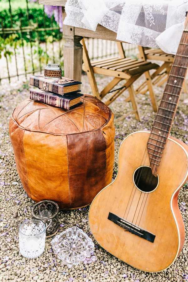 A styled decor vignette photographed outdoors on a gravel surface, featuring a tan leather Moroccan-style pouf topped with a stack of vintage hardcover books and a small decorative jar. An acoustic guitar leans against the pouf on the right side, and a wooden folding director's chair draped with white lace fabric is visible in the background. Small crystal and metal votive candle holders are arranged on the gravel in the foreground. The overall styling theme is bohemian with vintage touches, mixing leather, lace, books, and musical elements. Close-up/detail shot.
