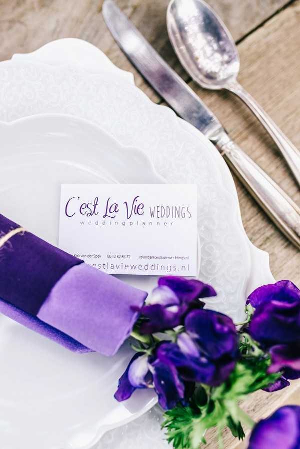 A close-up detail shot of a styled place setting featuring a white scallop-edged plate layered on a second white decorative plate, with a deep purple velvet ribbon-wrapped napkin roll and a small cluster of deep purple sweet pea flowers placed on top. A business card for 'C'est La Vie Weddings – weddingplanner' with the website cestlavieweddings.nl is propped against the napkin. Silver cutlery — a spoon and knife — rest to the upper right on a light wood surface. The color palette centers on white and deep violet-purple, creating a cohesive branded tablescape presentation.
