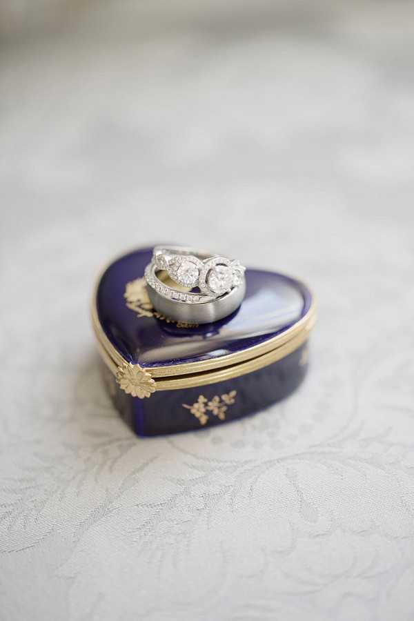 Close-up detail shot of wedding rings resting on top of a heart-shaped Limoges-style porcelain trinket box in deep cobalt blue with gold floral detailing and a gold clasp. The rings include a diamond engagement ring featuring a round solitaire with a pavé-set band, a diamond wedding band, and a plain silver-toned groom's band. The box is placed on a white textured linen or brocade surface. The composition is a tight macro shot with shallow depth of field, keeping the rings and box in sharp focus against a softly blurred background.