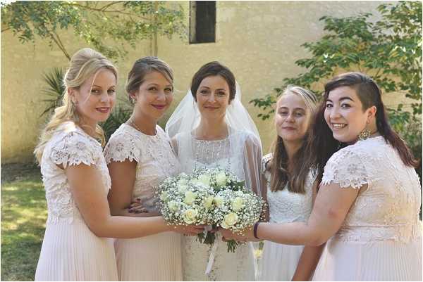 A portrait of a bride posing with four bridesmaids outdoors in front of a pale yellow stone building. The bride wears a white lace dress with a sheer veil and holds a bouquet of white roses and white baby's breath. The four bridesmaids are dressed in coordinating blush and ivory lace cap-sleeve dresses. The group is arranged in a close cluster facing the camera, with the bride at center. The shot is a mid-range portrait with a shallow depth of field, slightly blurring the building and garden greenery in the background.
