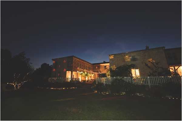 A wide-angle nighttime exterior shot of a rustic stone farmhouse or barn venue illuminated by warm amber interior lighting and string lights draped across the facade and surrounding trees. The venue features multiple interconnected stone buildings with lit windows, a white picket fence, and what appears to be an outdoor terrace or covered area strung with festoon lights. Small ground-level lights line a pathway in the foreground, creating a warm glow against the deep blue twilight sky. No people are visible in the frame. Potential venue feature image.