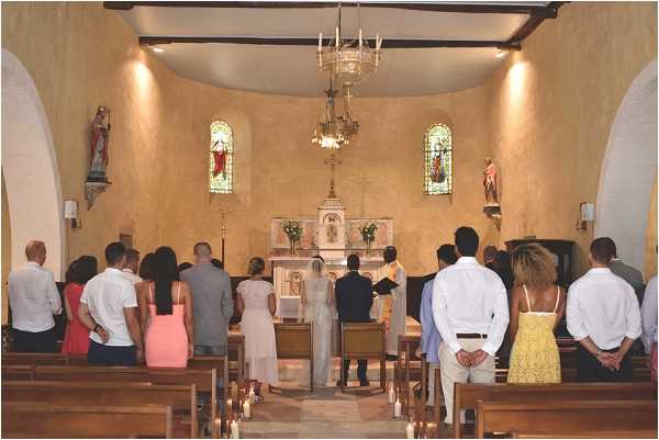 A wedding ceremony is taking place inside a small, intimate French chapel with warm ochre-toned plaster walls, dark wooden ceiling beams, and arched alcoves housing religious statues. The bride, dressed in a white or ivory gown, and the groom stand at the altar before an officiant, with the white marble altar decorated with green floral arrangements and candles. Approximately 15–20 guests stand in the wooden pews facing the altar, dressed in casual summer attire including a coral pink dress, a yellow sundress, and light-colored shirts. White pillar candles line the stone floor along the aisle, and a chandelier hangs above the nave, with stained glass windows in green and red providing color accents at the front of the chapel. This is a wide shot taken from the back of the chapel.