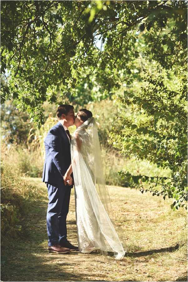 A couple shares a kiss during outdoor wedding portraits on a shaded path beneath overhanging tree branches. The groom wears a navy blue suit with brown leather shoes and a burgundy tie, while the bride wears a lace wedding gown with a long cathedral-length veil that trails behind her on the ground. The two are holding hands as they kiss, framed naturally by the tree canopy above. The shot is a full-length portrait taken in bright midday light with dappled shade.