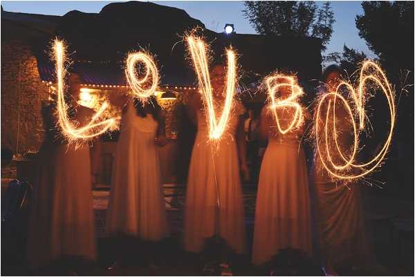 A long-exposure evening shot of five women in floor-length light-colored gowns standing outdoors and writing the word 'LOVE' (with an additional swirl) in the air using sparklers, creating glowing light trails against the dusk sky. The setting appears to be a rustic stone venue with warm string lights visible in the background. The women are likely bridesmaids or members of the bridal party, all dressed in matching or coordinating flowing dresses. The composition is a wide portrait shot with the sparkler light trails as the focal point.