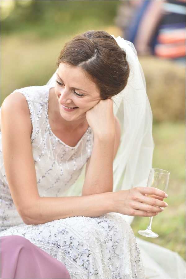 A close-up portrait of a bride sitting outdoors, leaning her head on her hand in a relaxed pose while holding a champagne flute. She is wearing a sleeveless, heavily sequined and beaded ivory wedding dress with a long white veil, her dark brown hair styled in an updo. The background is softly blurred and appears to be an outdoor field or garden setting, with what looks like a hay bale and a guest in the far background wearing a mauve/purple outfit. The image has a candid, unposed quality captured during what appears to be a cocktail hour or outdoor reception.