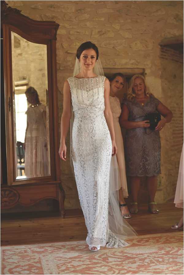 A bride is shown getting ready indoors in what appears to be a rustic chateau or stone-walled room, standing on a terracotta and cream patterned rug. She wears a fitted, sleeveless, floor-length gown heavily embellished with silver beading and lace detailing throughout, paired with a sheer veil and white open-toe heels. Her hair is styled in an updo. Behind her, two women — one in a blush/nude dress and one in a silver-grey lace knee-length dress — look on admiringly, and a large ornate wooden mirror to the left reflects the bride's back. The composition is a full-length portrait shot taken indoors with warm natural light.