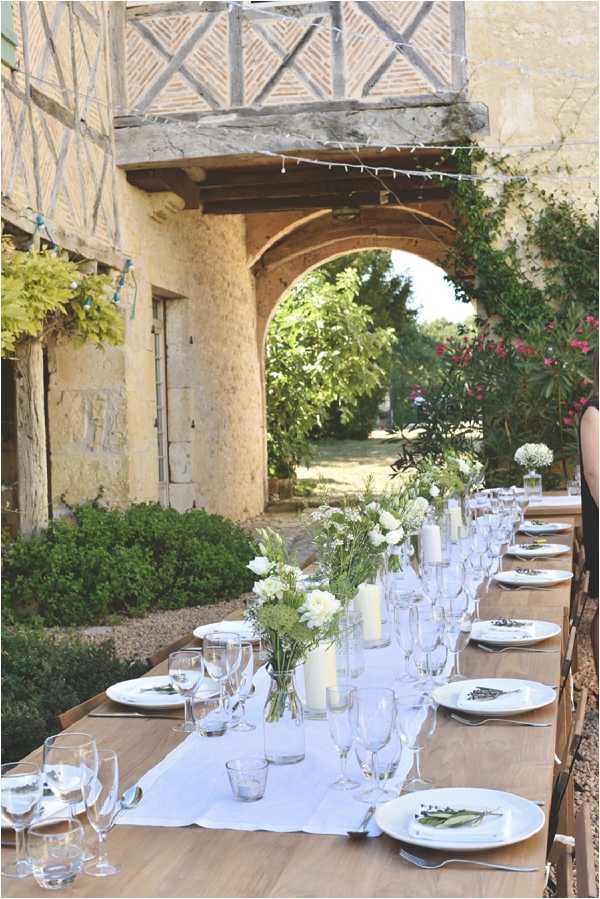 An outdoor wedding reception table is set for a long guest dinner alongside a historic French manor or farmhouse with exposed timber framing, stone arches, and a covered gallery overhead strung with small bunting or fairy lights. The long wooden farm table is dressed with a white linen runner and set with white ceramic plates, multiple wine and champagne glasses, and silver cutlery. Centerpieces consist of clusters of small glass bud vases and cylinder vases holding white ranunculus, Queen Anne's lace, and wild greenery, interspersed with ivory pillar candles of varying heights. The overall decor palette is white and natural wood, giving a rustic yet clean aesthetic. A single guest in a dark dress is partially visible at the far right edge. The shot is a wide portrait-format image taken from the near end of the table looking down its full length toward the archway and garden beyond. Potential venue feature image.