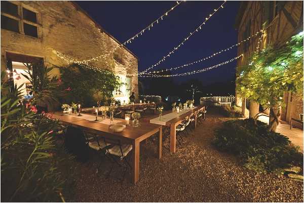 An outdoor evening wedding reception set in a courtyard or garden area adjacent to a stone building, photographed as a wide shot. Multiple long wooden trestle tables with folding chairs are arranged on a gravel surface, dressed with white floral centerpieces, candles, and glassware in a relaxed, rustic style. Strings of warm Edison/fairy lights are strung overhead in a canopy pattern, providing the primary lighting along with candles on the tables. Lush potted plants and climbing greenery border the space. No guests are visible, suggesting the photo was taken before guest arrival. Potential venue feature image.