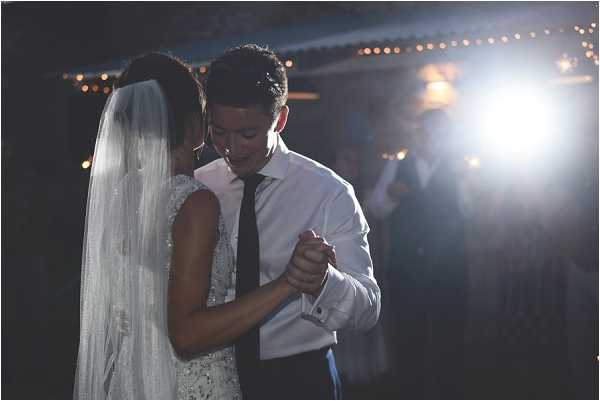 The bride and groom share their first dance in a darkened indoor reception space, lit by a single bright spotlight and warm fairy lights strung overhead in the background. The bride wears a fitted lace or beaded gown with a long white veil, while the groom is dressed in a white dress shirt with a dark tie and dark trousers, his jacket removed. The couple holds hands and dances close together, with the groom looking downward in an intimate moment. The image is a mid-shot portrait with a dark, moody atmosphere created by the low ambient lighting and strong directional spotlight.