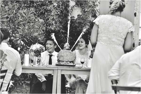 A black-and-white image of a wedding reception moment, likely a cake cutting or dessert presentation, captured outdoors or in a garden-adjacent space with dense foliage visible in the background. The couple is seated at a wooden sweetheart table; the groom wears a white dress shirt and dark tie, while the bride wears a lace-detailed dress and veil, both smiling and holding lit sparklers that are actively sparkling above a decorated cake or dessert platter on the table. A small floral arrangement, candles, and wine glasses are visible on the table. In the foreground, a partially visible figure in a lace-back wedding gown — likely a bridesmaid or second bride — is turned away from camera. The image is shot in a medium wide candid style with soft contrast and bright highlights from the sparklers.