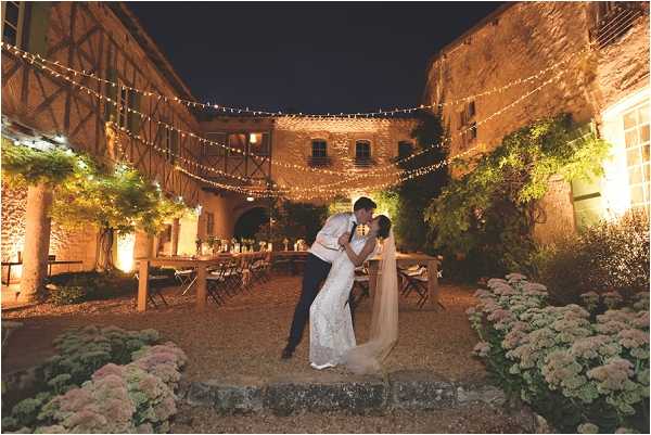 A couple shares a dip kiss in the foreground of an outdoor courtyard reception at night, with the groom in a dark suit and the bride wearing a fitted lace gown with a long veil. The courtyard of what appears to be a historic stone and half-timbered venue is strung with multiple lines of warm fairy lights overhead, creating a canopy effect across the space. Behind the couple, long wooden farm-style dining tables with folding chairs are set up for the reception, lit by warm uplighting on the stone walls and surrounded by flowering plants in pale pink tones. Wide-angle portrait shot capturing both the couple in the foreground and the full venue setting. Potential venue feature image.