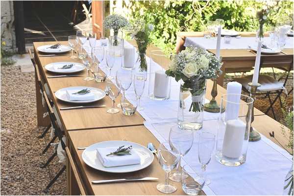 A wide-angle detail shot of an outdoor wedding reception table set on a gravel surface, likely a courtyard or garden terrace. The long wooden farm-style table is dressed with a white linen runner and set with white plates, folded white napkins garnished with small sprigs of lavender, and clear crystal champagne flutes and wine glasses. Centerpieces include low glass vases with white roses and baby's breath, alongside tall white pillar candles and slender taper candles in brass and glass holders. The overall decor palette is white and green with rustic-classic styling. Black folding chairs are partially visible along the sides of the table.