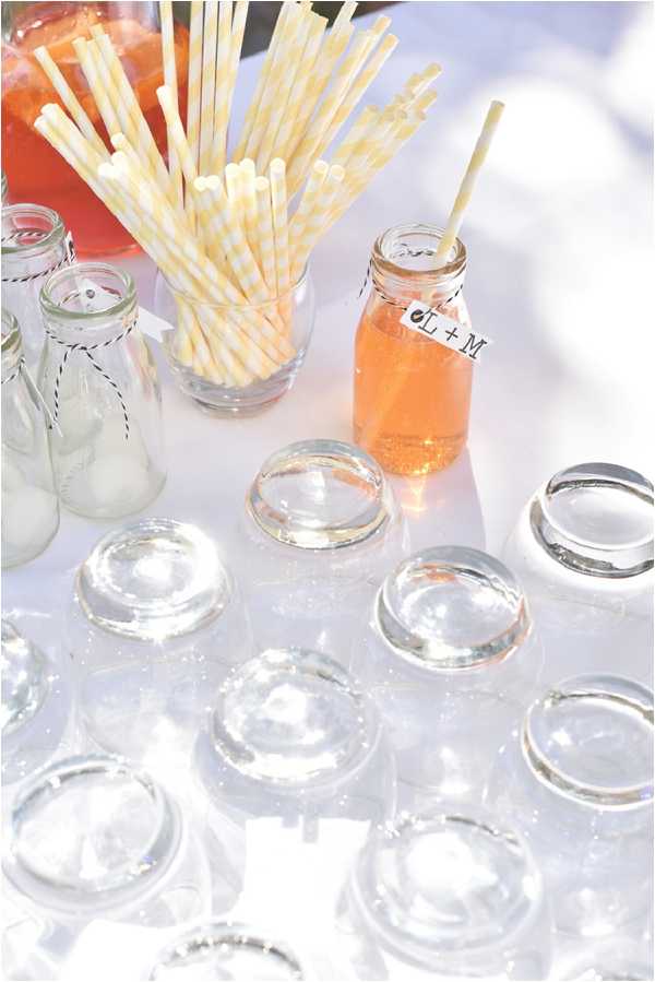 Close-up detail shot of a drinks station at an outdoor wedding, set on a white linen-covered table in bright sunlight. Several upside-down clear glass tumblers are arranged in the foreground, alongside small vintage-style glass milk bottles tied with black-and-white baker's twine. In the background, a glass jar holds an assortment of yellow and white striped paper straws, and a small mason jar filled with an amber-orange drink is labeled with a handwritten tag reading 'L + M,' indicating the couple's initials. The styling is casual and rustic, consistent with a DIY or bohemian aesthetic.