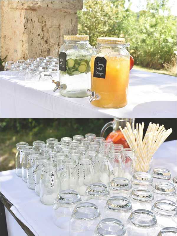Detail shots of an outdoor cocktail hour drinks station at a French wedding, shown in two panels. The top image features two large glass beverage dispensers on a white-clothed table — one containing cucumber-infused water labeled 'Gin Tonic' and one containing an orange juice or peach-colored drink labeled 'Harvey Wall Banger' — with small chalkboard tags and gold lids, surrounded by rows of empty glass tumblers. The bottom image shows a matching drinks table with multiple small personalized glass milk bottles labeled with what appears to be initials 'N + M' tied with black-and-white twine, alongside mason jar-style glasses and a bundle of yellow-and-white striped paper straws in a glass holder. The overall styling is rustic-casual with a yellow and white color palette.