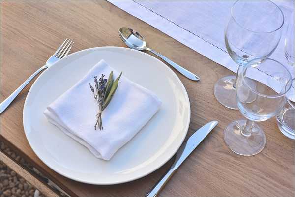 A close-up detail shot of a single place setting on a natural wood dining table. A white ceramic plate holds a neatly folded white linen napkin tied with a small sprig of lavender and an olive leaf as a napkin accent. Silver flatware — a fork, knife, and spoon — is arranged around the plate, and two clear wine glasses are positioned to the right. A white hemstitch table runner is partially visible at the top of the frame. The overall decor palette is white and natural wood with a Provençal-inspired rustic styling.