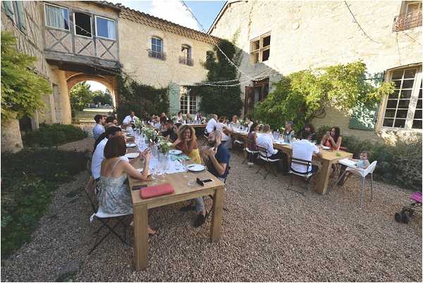 An outdoor wedding reception dinner is taking place in a gravel courtyard of a rustic French stone farmhouse or mas, with timber-framed building elements and ivy-covered walls visible in the background. Approximately 30–40 guests are seated along several long wooden tables arranged in a loose U-shape, with white chairs and folding black chairs mixed throughout. The tables are set with white plates, glassware, and low centerpieces of greenery and white flowers, with string lights strung overhead between the buildings. The scene is captured in a wide-angle overhead shot, conveying the full scale of the outdoor dining setup in an informal, relaxed rustic style. Potential venue feature image.