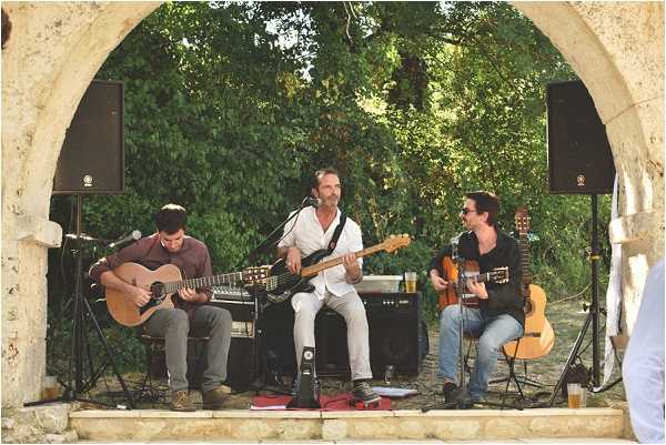 Three male musicians perform live during what appears to be a wedding reception cocktail hour or outdoor entertainment set. They are positioned under a large stone arch, with two playing acoustic guitars and one standing playing an electric bass guitar at a microphone. The performers are dressed casually in jeans and light shirts. Black Yamaha speaker cabinets are placed on either side of the arch, and audio equipment including a mixing board is visible on a table behind the bassist. The setting is an outdoor stone structure, likely part of a French chateau or historic venue, with green trees visible through and behind the arch. Wide shot captures the full performance space and surrounding stonework.