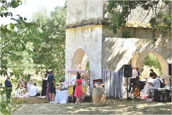 A wedding cocktail hour taking place outdoors beneath a series of stone arches, likely part of a historic aqueduct or ruined arcade structure. Approximately 15 guests are mingling in small groups, with some seated on hay bales and others standing near white-clothed cocktail tables. A vintage-style yellow and white cart serves as a drinks or candy station, positioned next to a wooden wine barrel. On the right side, musicians are set up with instruments and a PA speaker, suggesting live music entertainment. The decor style is rustic and relaxed, incorporating natural materials like hay bales and barrels. The shot is a wide medium-distance image capturing the full scene.