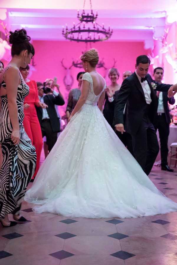 The bride and groom are dancing at their indoor wedding reception, surrounded by guests who are also dancing and watching. The bride wears a full ballgown with a lace-appliquéd tulle skirt, cap sleeves, and a low open back, with her hair in an updo; the groom is in a dark suit with a black bow tie and a white boutonniere. The ballroom walls are lit with vivid magenta/hot pink uplighting, and decorative stag antler mounts are visible on the walls alongside a wrought-iron chandelier overhead. The floor features a geometric tile pattern, and guests in the background are dressed in varied formal attire including a black-and-white zebra-print gown and a coral/red dress; the shot is taken from behind the couple at a medium distance, capturing the full sweep of the bridal gown.