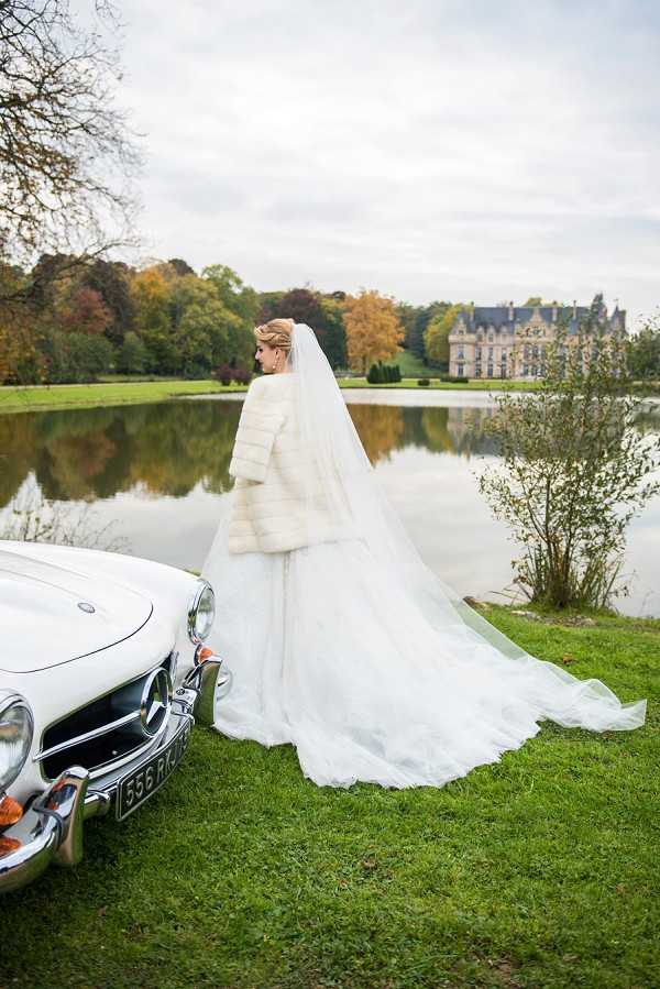 A bride portrait taken outdoors on the grounds of a French chateau, shot from behind in a wide portrait composition. The bride wears a full-length white ballgown with a long cathedral veil and a white fur bolero jacket, her hair pinned up with a decorative accessory. She stands beside a vintage white Mercedes-Benz convertible on a lawn at the edge of a reflective lake, with the chateau's grand facade visible across the water in the background. The setting and classic vehicle give the image a formal, vintage-inspired aesthetic, photographed during autumn as indicated by the orange and yellow foliage on surrounding trees. Potential venue feature image.