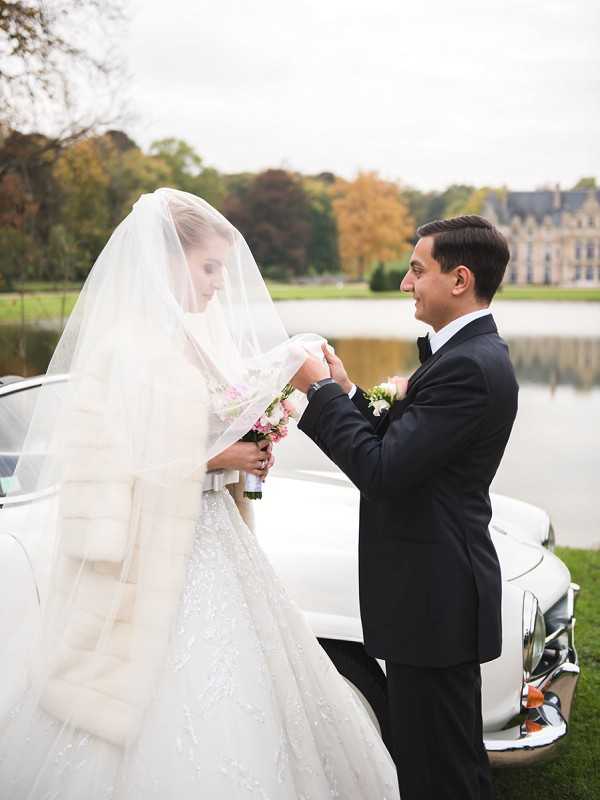 A couple portrait taken outdoors on the grounds of a French chateau, visible in the background across a reflective pond. The groom, wearing a black tuxedo with a white boutonniere, lifts the bride's long cathedral-length veil as she looks down. The bride wears a heavily embellished ball gown with a white fur stole wrap and carries a small bouquet of blush and white florals. A classic white vintage car is partially visible behind them. The styling is formal and classic. Medium portrait shot.