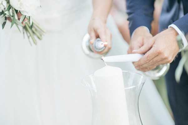 Close-up detail shot of a unity candle ceremony, showing the couple's hands lighting a large white pillar candle housed in a clear glass hurricane vase. The bride, visible in a white dress and holding a bouquet of white blooms and eucalyptus, holds one taper candle on a silver holder, while the groom, wearing a navy suit, holds the other. The composition is tightly framed on the hands and candle, with the background softly blurred.