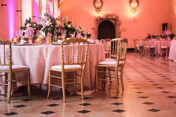 A wide shot of an indoor wedding reception setup inside a ballroom with ornate architecture, pink-washed walls, and a black-and-white diamond-patterned marble floor. The foreground features a round table dressed in a blush pink floor-length linen, surrounded by gold Chiavari chairs with ivory cushions, and a lush centerpiece of blush pink hydrangeas, dusty mauve and ivory blooms, and dark greenery in gold vessels alongside taper candles. In the background, long rectangular guest tables are similarly styled with blush linens and floral arrangements, and the room is bathed in pink ambient uplighting. The overall decor palette is blush, gold, and mauve with a classic, formal styling theme. Potential venue feature image.
