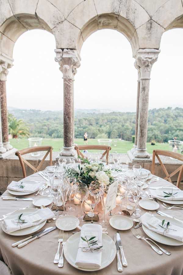 A reception table detail shot taken on an open stone arcade loggia with Romanesque arched columns, overlooking a wide landscaped grounds below. The round table is dressed with a taupe linen tablecloth and set for approximately eight guests, with white china, silver cutlery, and multiple crystal wine and champagne glasses per place setting. Each place setting includes a white linen napkin folded and tied with twine around a small olive branch sprig. The centerpiece is a low arrangement of white peonies and lush greenery, surrounded by small lit votive candles. The overall decor palette is neutral and white with green botanical accents, giving a classic French countryside feel. Potential venue feature image.