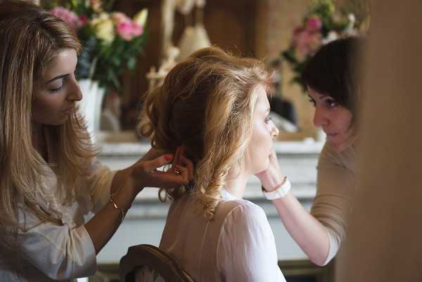A getting-ready scene showing a bride seated while two women work on her hair and makeup simultaneously. The setting appears to be an interior room with an ornate fireplace mantel visible in the background, decorated with arrangements of pink and cream flowers in white vases. The bride has auburn hair styled in a loose updo with soft curls, and is wearing a pale blush or ivory top. The woman on the left, with long blonde hair, is adjusting the bride's hair, while the woman on the right, with short dark hair, attends to her face. The image is a medium close-up portrait shot with a shallow depth of field, slightly soft focus in the background.