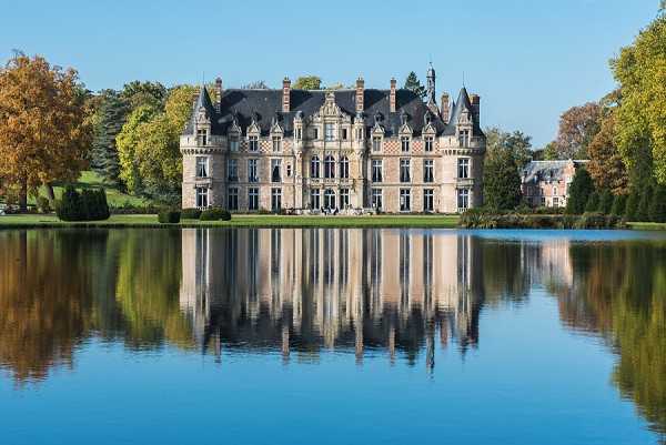 Wide exterior shot of a French chateau with classic 19th-century architecture, featuring ornate stonework, steep slate mansard roofs, corner turrets, and multiple chimneys. The building is reflected almost perfectly in a large ornamental lake in the foreground, shot on a clear day with autumn foliage visible in the surrounding trees in shades of gold and green. No people are visible in the frame. Potential venue feature image.