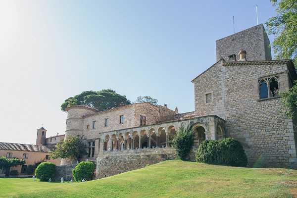Wide exterior shot of a historic stone château or castle-like venue photographed in bright daylight. The building features medieval-style architecture including a prominent square tower, an arched stone colonnade gallery running along the facade, and rounded stone structures typical of southern French or Catalan heritage buildings. The grounds include manicured topiary shrubs and a sloped lawn in the foreground. No people, wedding party, or decor elements are visible in this image. Potential venue feature image.