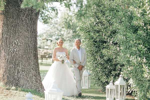 A bride walks down an outdoor aisle accompanied by a man in a light beige suit, likely her father, during a garden ceremony. The bride wears a full-skirted white ballgown and carries a white bouquet, with her blonde hair styled up. The aisle is lined with white freestanding lanterns of varying heights, and the path is framed by large trimmed hedges and a broad tree trunk, creating a natural corridor. The composition is a medium wide shot taken at ground level, capturing the processional in a classic, garden-style setting.
