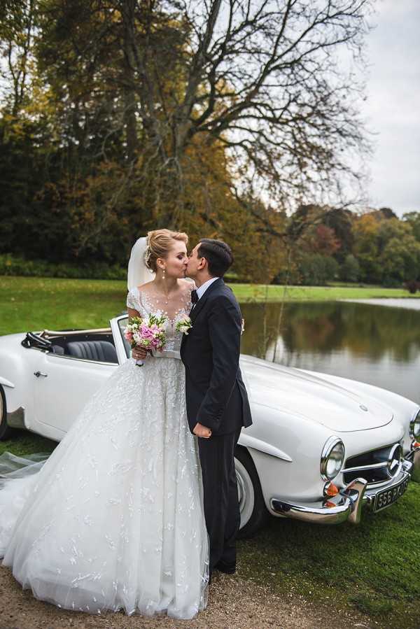 A couple portrait taken outdoors on the grounds of what appears to be a French estate or chateau, with a pond and mature trees visible in the background. The bride and groom are kissing in front of a white vintage Mercedes-Benz 190SL convertible. The bride wears a full-length ballgown with a lace and floral-embroidered tulle skirt, an illusion neckline bodice, short sleeves, and a long cathedral veil styled in an updo; she holds a bouquet of pink peonies and small white blooms. The groom wears a classic dark navy suit. The shot is a medium full-length portrait with the vintage car serving as a prominent prop in a classic, romantic wedding style.