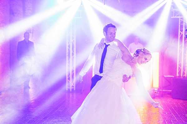 A couple performs their first dance on an indoor dance floor surrounded by dramatic stage lighting and artificial fog or dry ice effects. The groom wears a white dress shirt with a navy blue tie and dark trousers, while the bride wears a full-skirted white ballgown with her blonde hair styled up. Multiple beam lights mounted on metal trussing cast purple, pink, and white rays across the scene, creating a concert-like atmosphere. A few guests or event staff are visible in the background. The shot is a mid-range portrait capturing the couple in motion, with the groom dipping or spinning the bride who is laughing and leaning back.
