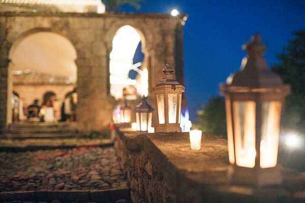 An outdoor evening wedding reception scene photographed with a shallow depth of field, focusing on a row of white metal lanterns and small votive candles lining a stone balustrade or terrace wall. In the soft-focus background, guests are gathered beneath large stone archways of what appears to be a historic chateau or manor, warmly lit from within against a deep blue twilight sky. The lanterns cast a warm amber glow along the stone ledge, creating a pathway of light leading toward the venue entrance. The decor style is classic and romantic, relying entirely on candlelight and lantern lighting with no visible floral elements. Potential venue feature image.