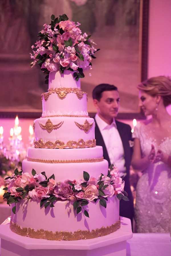 A close-up shot of a large five-tier wedding cake displayed during an indoor reception, bathed in pink ambient lighting. The cake is white fondant with ornate gold baroque-style relief decorations on each tier, including scrollwork, medallions, and swag details; the base and upper tiers are adorned with clusters of pink and mauve sugar or fresh roses with green foliage, and the top is crowned with a large arrangement of pink blooms. In the soft background, a couple — the groom in a dark suit and the bride in a fitted, heavily beaded or sequined ivory gown — stand together conversing, flanked by lit taper candles on candelabras. The reception setting appears to be an ornate indoor ballroom or chateau salon with warm candlelight and pink uplighting creating a rosy glow throughout.