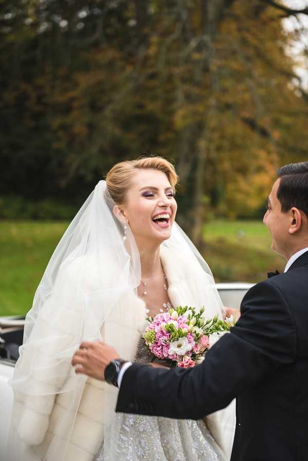 A bride and groom share a candid, joyful moment outdoors, with the bride laughing openly as the groom holds her by the waist. The bride wears a beaded or lace gown with a white fur stole and a long white veil, styled with an updo and bold purple eye makeup. She holds a bouquet of pink spray roses, white lisianthus, and green accents. The groom is dressed in a black tuxedo with a bow tie. A vintage white car is partially visible in the background. The portrait is a medium close-up shot taken in natural outdoor light during an autumn setting.