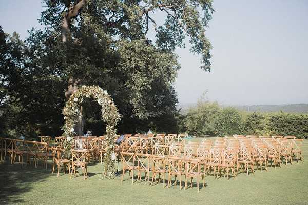 An outdoor ceremony setup on a lawn, with rows of natural wood cross-back chairs arranged on either side of a central aisle. A circular floral arch serves as the focal point at the altar, decorated with greenery and small white flowers. The setting has an open, panoramic backdrop with a large tree providing shade and a distant landscape visible beyond the grounds. The overall decor style is rustic-natural, using warm wood tones and an organic greenery-forward palette. Wide shot capturing the full ceremony layout. Potential venue feature image.