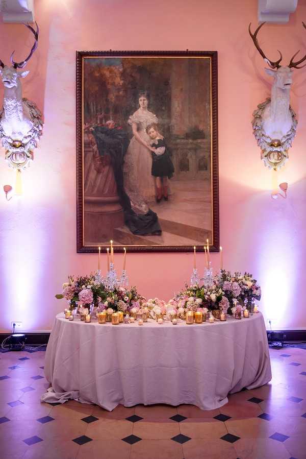 Indoor reception detail shot of a sweetheart or head table inside what appears to be a chateau ballroom or grand salon. The round table is draped in a pale grey linen and decorated with an abundant arrangement of blush pink, mauve, and white flowers including garden roses and hydrangeas, flanked by tall gold candelabras with lit taper candles and scattered gold votive candle holders. The table is positioned directly beneath a large oil painting in a gilt-edged frame depicting a woman in a Victorian-era white gown with a child, hung on a warm pink-toned wall. On either side of the painting are mounted white painted stag heads with ornate decorative brackets, and soft purple uplighting illuminates the wall on both sides. The floor features a classic geometric tile pattern in terracotta and dark grey. The overall decor palette combines blush pink, gold, and grey in a classic, chateau-appropriate style. Wide shot capturing the full table and wall backdrop.