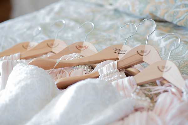 A detail close-up shot taken during a bridal getting-ready scene, showing several personalized wooden hangers laid out on a bed with light blue and cream paisley bedding. The hangers feature laser-engraved names and decorative motifs including arrows and floral wreaths, with one reading 'Marion' clearly visible. A white textured wedding dress is draped in the foreground alongside blush pink fabric pieces and pearl jewelry. The styling reflects a classic, feminine aesthetic with a blush and white color palette.